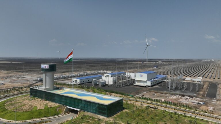 Aerial view of Adani Green's new battery storage facility in Rajasthan, showing solar panels and modular lithium-ion battery units with high-voltage connection lines.