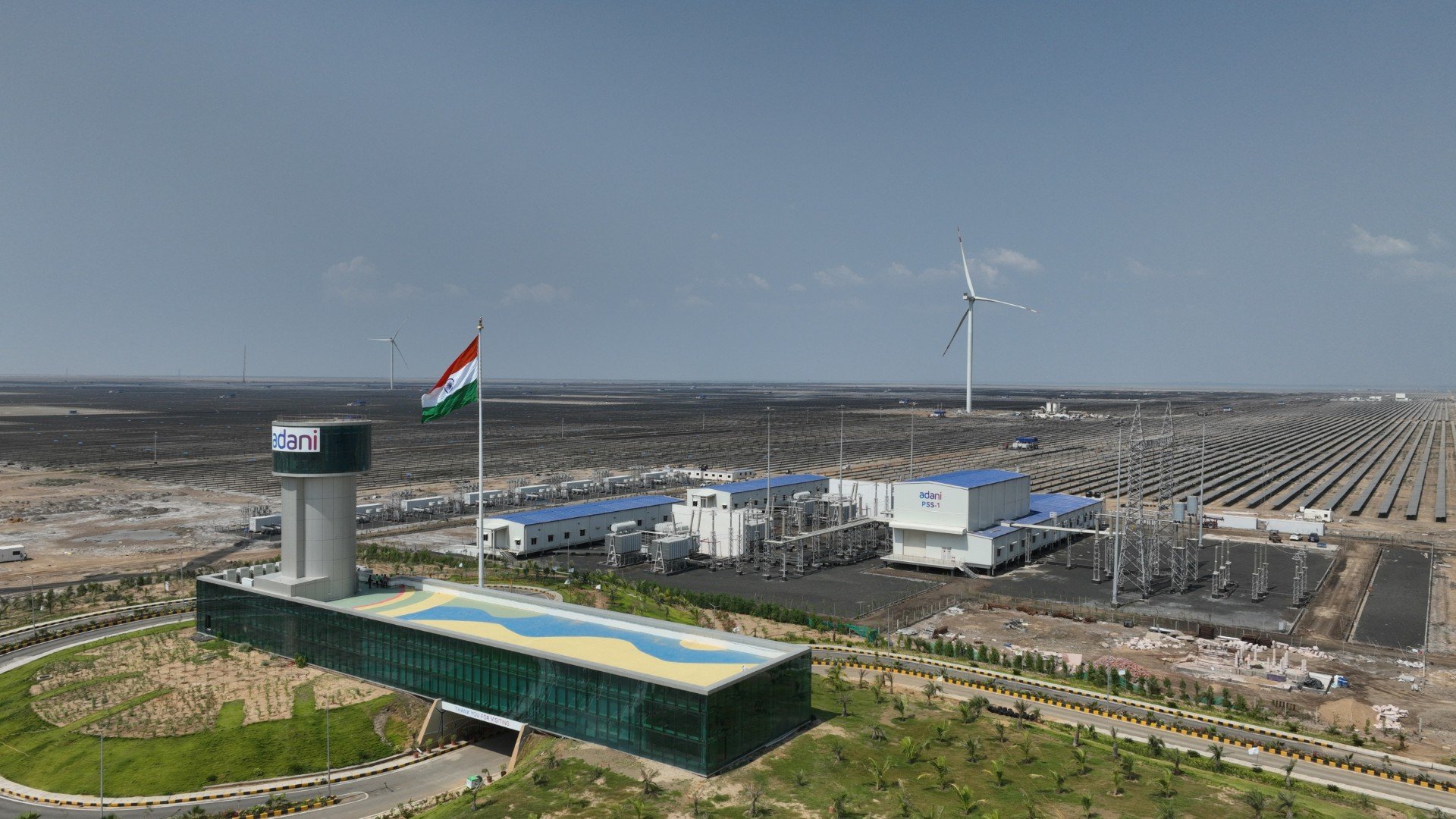 Aerial view of Adani Green's new battery storage facility in Rajasthan, showing solar panels and modular lithium-ion battery units with high-voltage connection lines.
